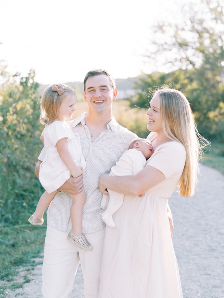 Young family smiling at each other, standing on a gravel path outside holding a toddler and newborn baby at their photoshoot with a family photographer in crestwood, ky